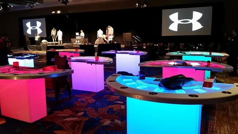 Glow casino tables lit pink and teal at a branded corporate event with stage screens